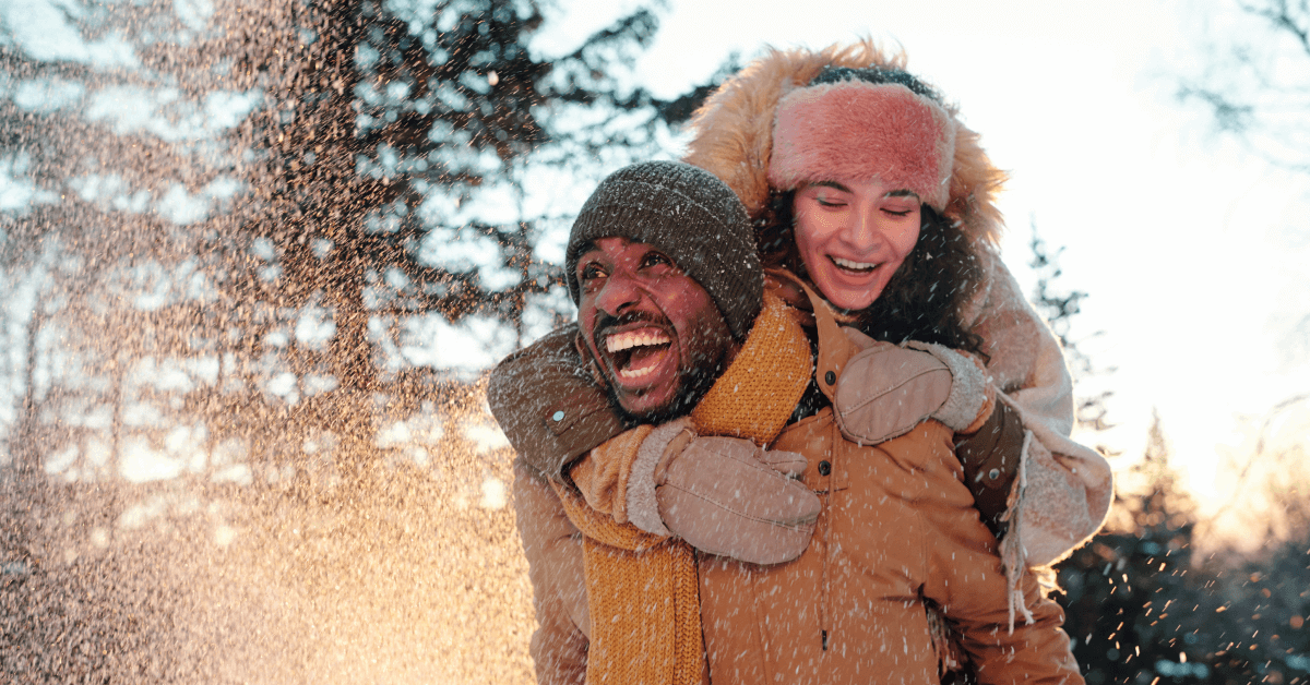 a man smiles while holding a woman on his back and snow hangs in the air
