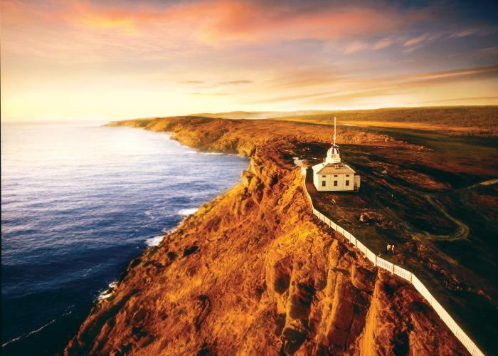 An aerial shot at sunrise shows a white picket fence along a rocky red cliff at the Cape Spear National Historic lighthouse. 