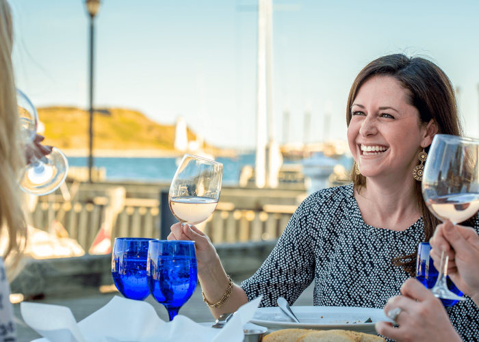 A smiling brunette woman raises a glass of white wine during a patio dinner at a restaurant on the Halifax waterfront. 