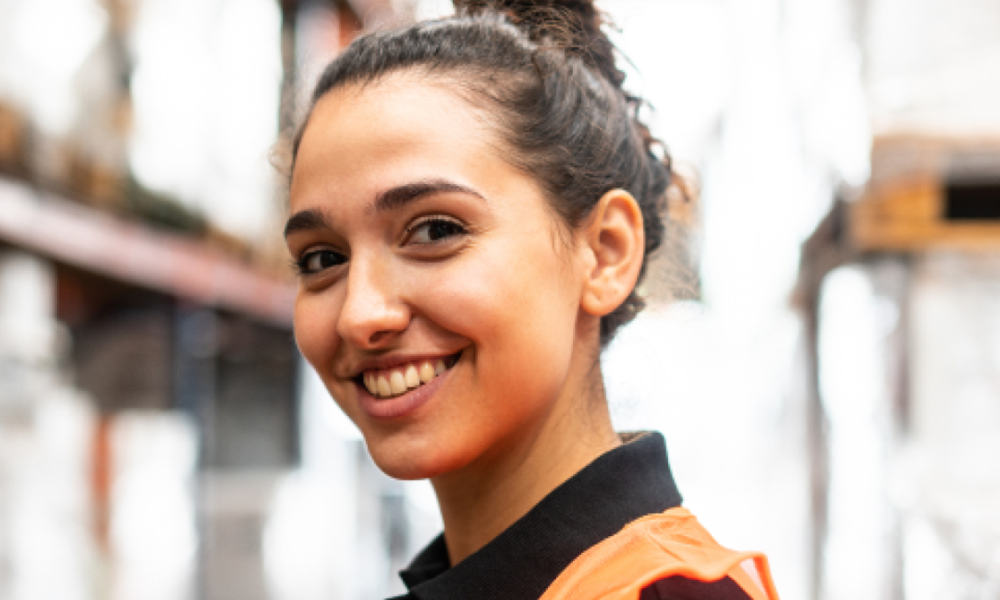 A smiling young person with their hair up, wearing an orange high-visibility vest and glancing over their shoulder.