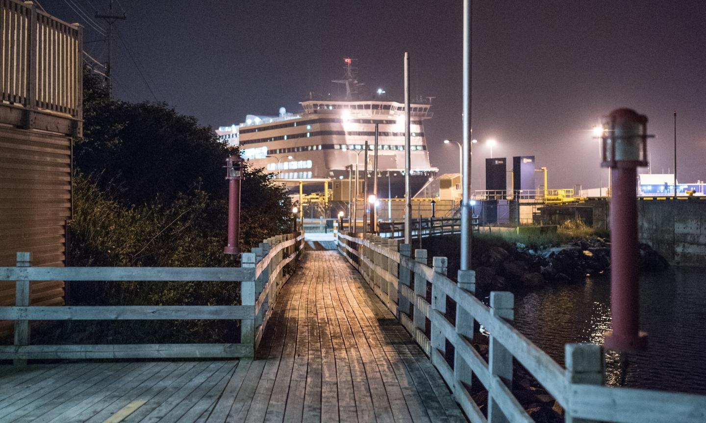 A wooden boardwalk leading toward a ferry terminal at night, with a large vessel illuminated against a dark, hazy sky.