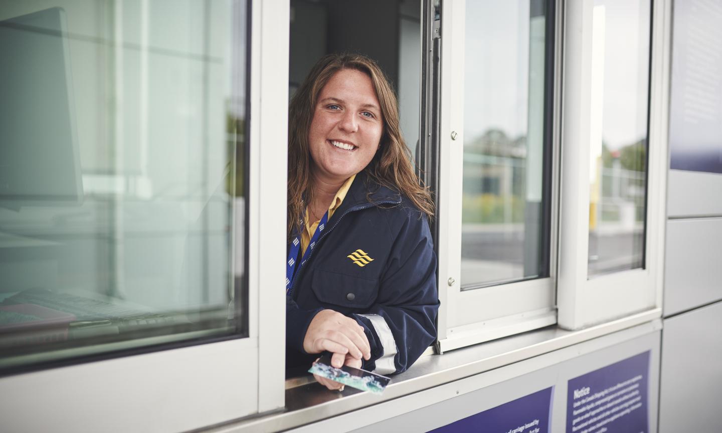 A smiling ferry employee in a navy uniform leaning out of a ticket booth window, holding a boarding pass.