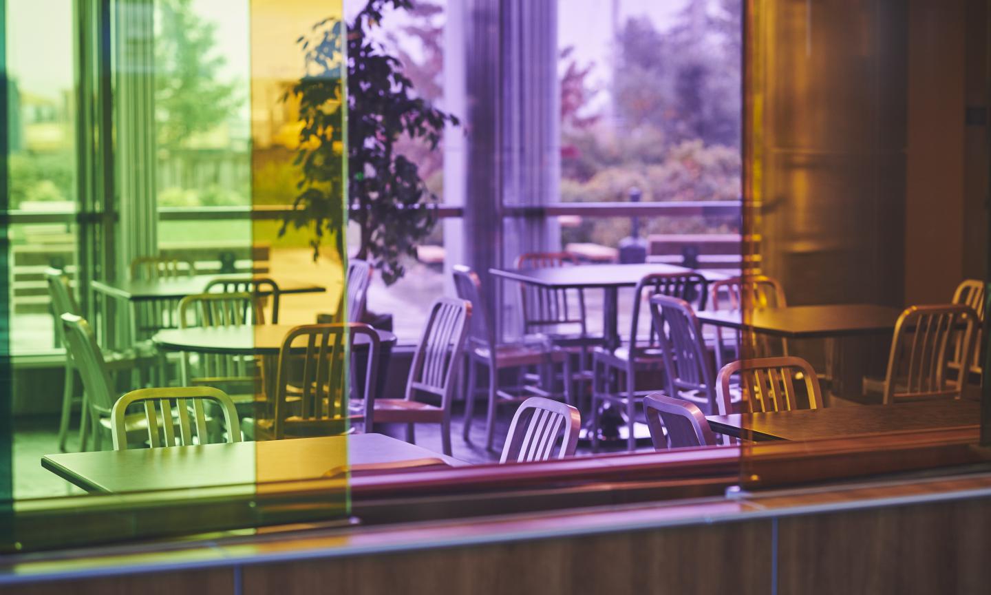 A restaurant dining area viewed through colourful tinted glass panels in green, yellow, purple, and orange, with trees visible through large windows beyond.