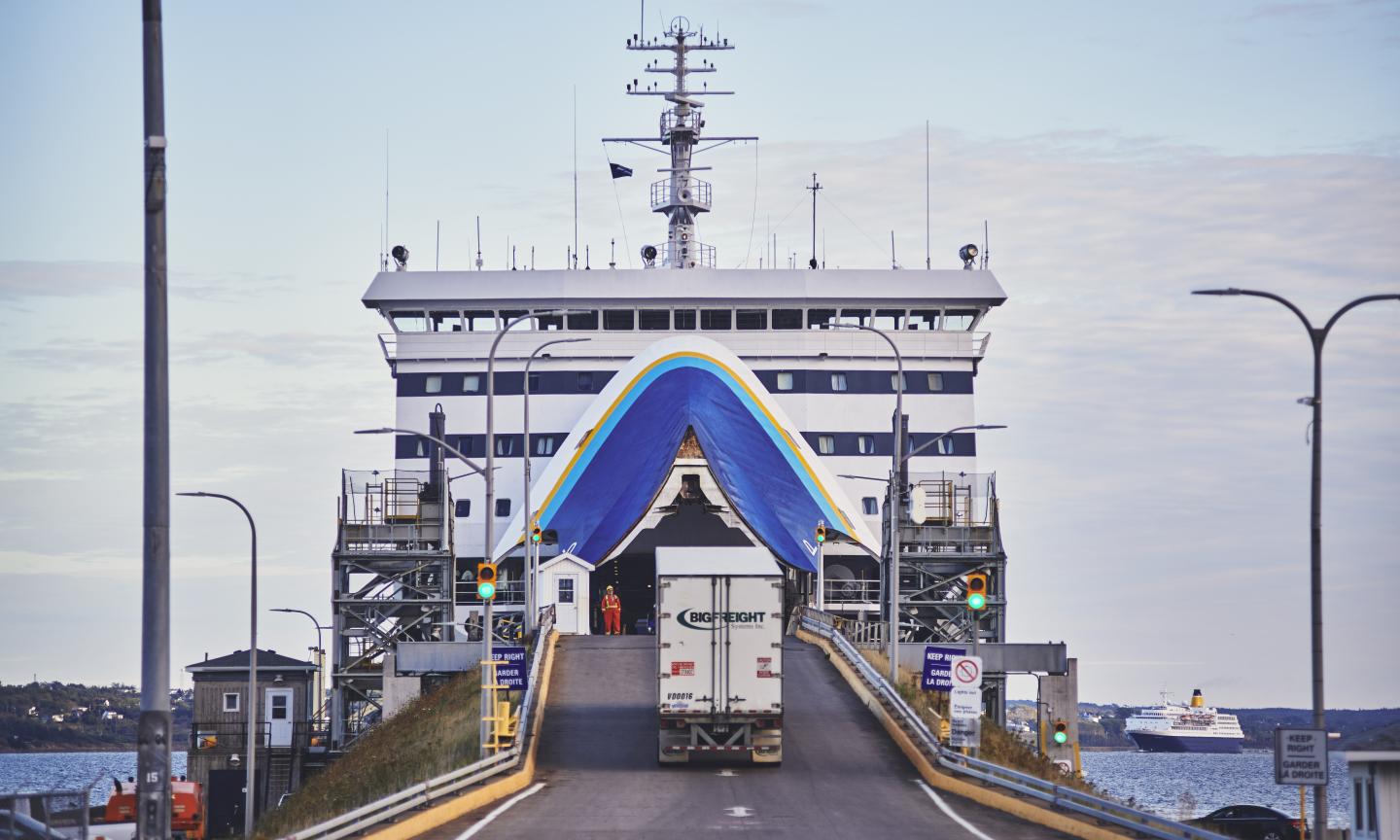A transport truck driving up a ramp into the open bow of a ferry, with a port worker in high-visibility gear standing nearby.