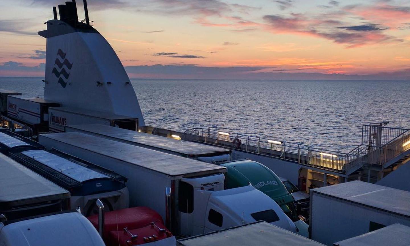 Rows of transport trucks parked on the open car deck of a ferry at sunset, with calm open ocean and a pink and orange sky stretching to the horizon.