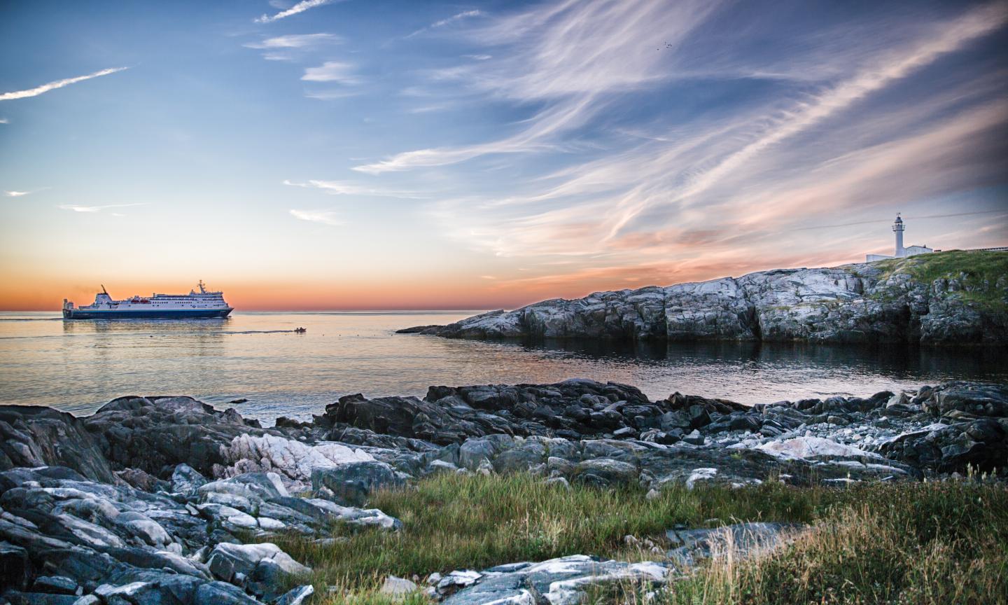 A ferry crossing calm waters at sunset past a rocky coastline with a white lighthouse, under a sky of pink and blue.