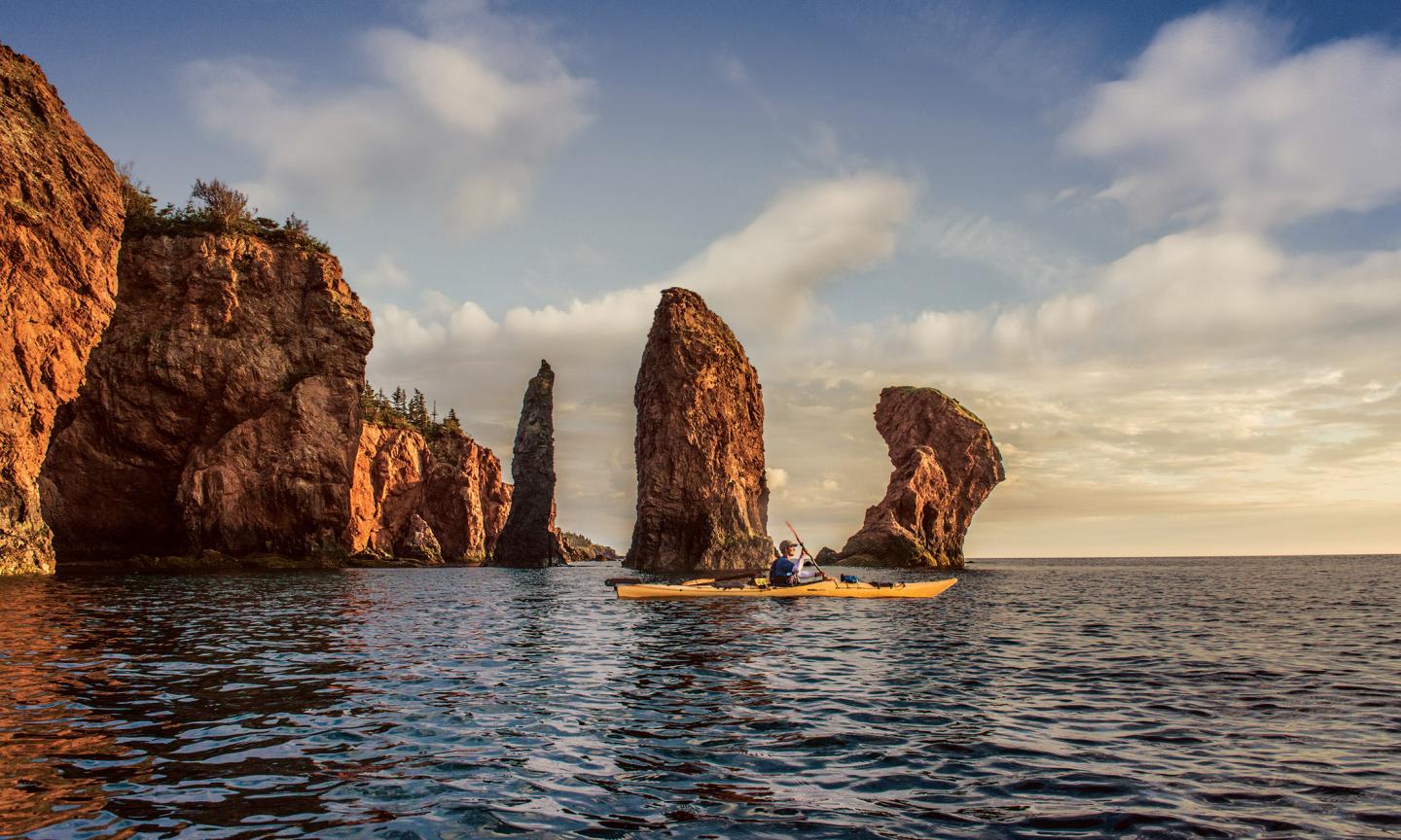 A kayaker paddling among red sandstone sea stacks rising from dark reflective water in warm golden light.