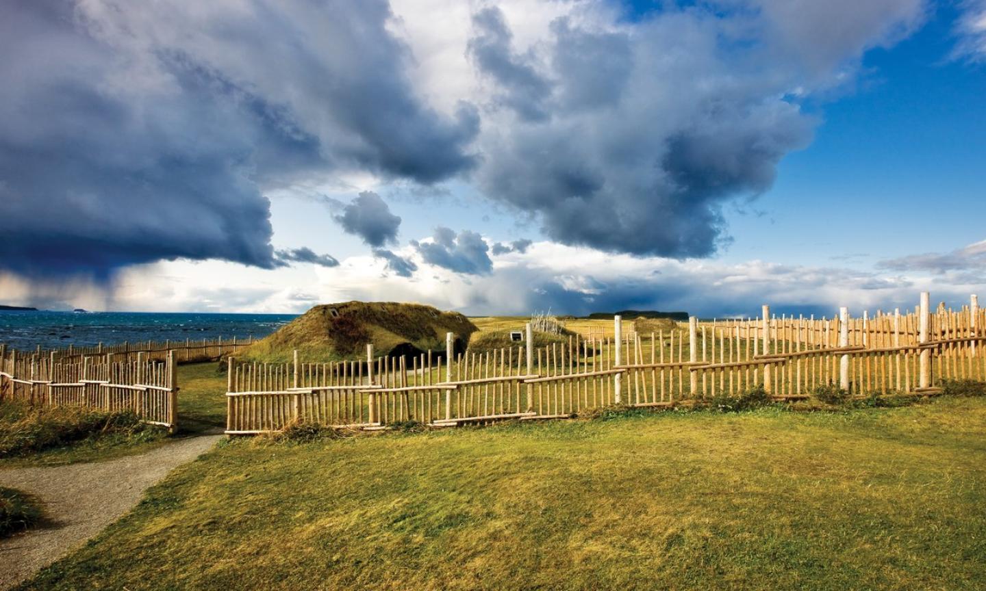 A sod-roof longhouse behind a rustic wooden palisade fence on a coastal plain.
