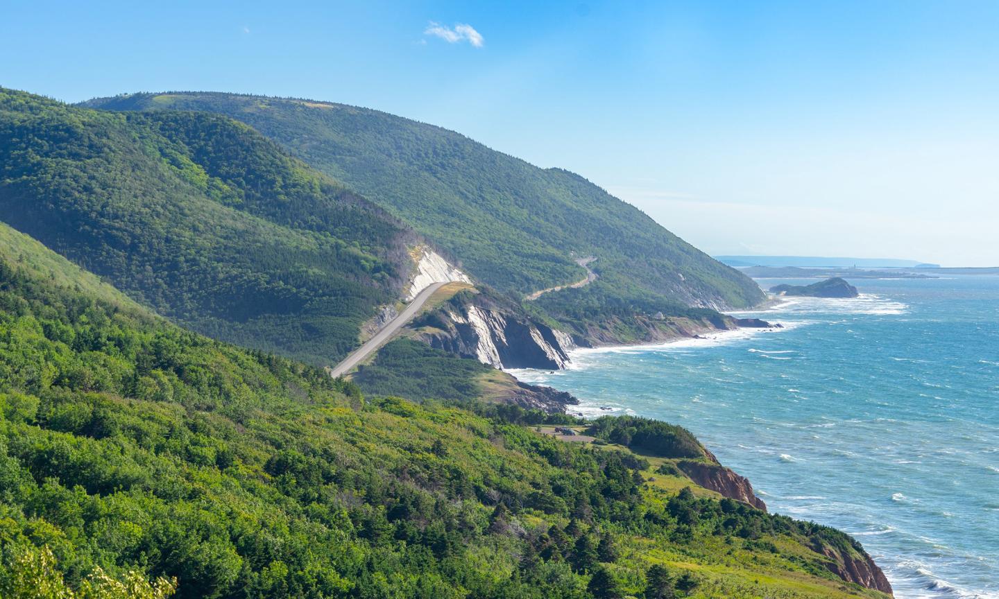 A scenic coastal highway carved into a steep clifftop, winding along a rugged shoreline of forested hills and crashing waves.