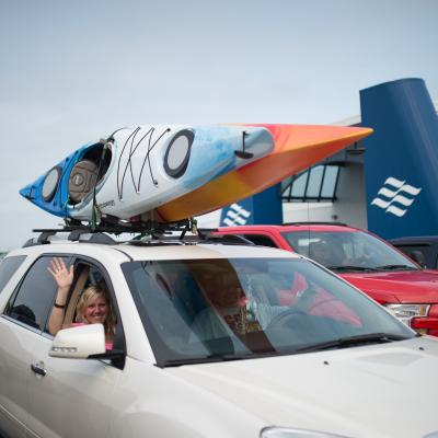 Passenger waving from car with kayaks on roof.