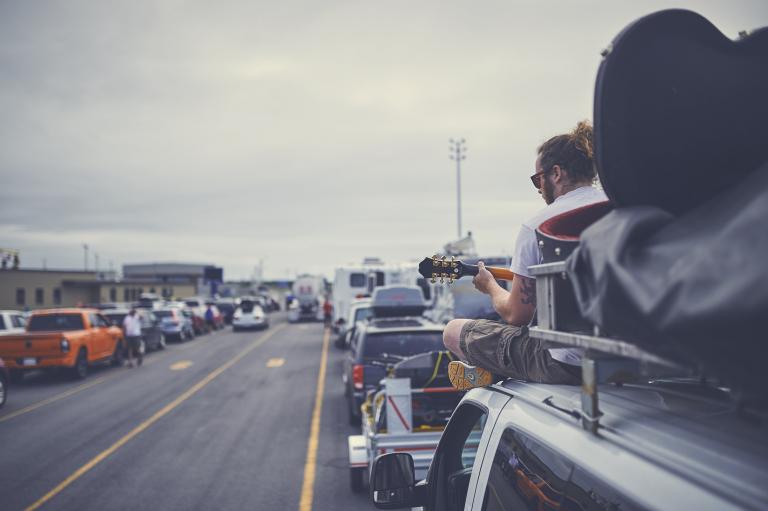 A ponytailed man in his twenties sits on top of a van and plays guitar while waiting to board a Marine Atlantic ferry. 