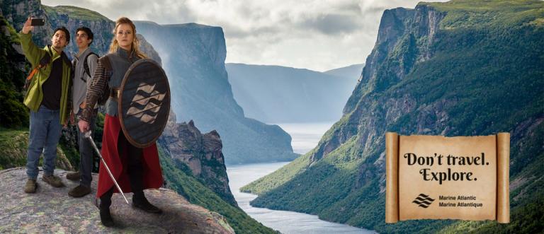 Image of a couple taking a selfie with Gros Morne in the background and a female viking in front. Text: Don't Travel. Explore