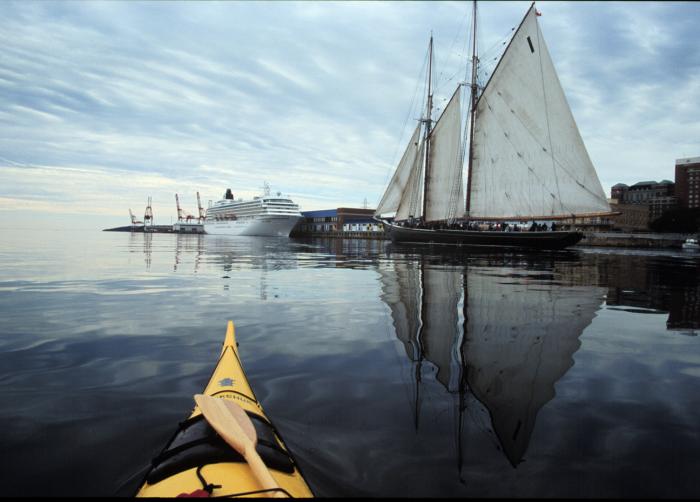 A yellow tip of a kayak on still water points toward Bluenose and cruise ship docked in Halifax port