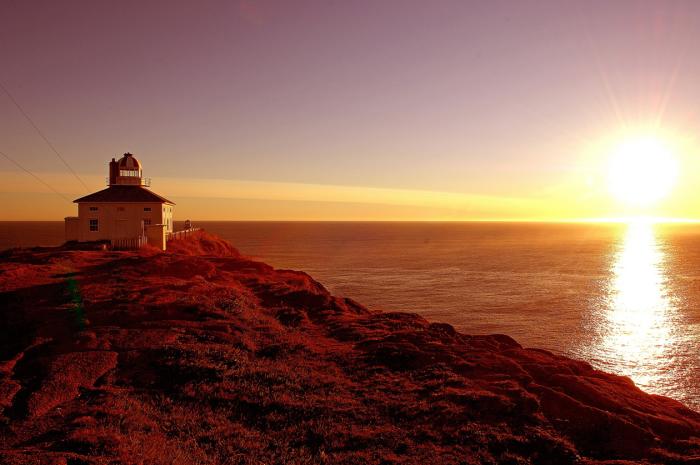 lighthouse on the edge of a cliff beside the ocean with sun rising in background