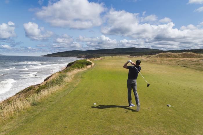 person swings golf club behind them on green with ocean's edge on their left