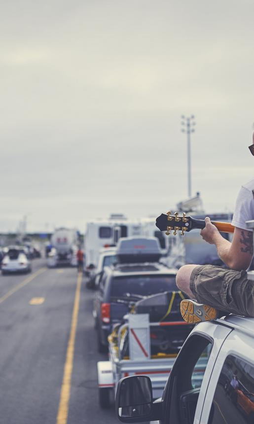 A ponytailed man in his twenties sits on top of a van and plays guitar while waiting to board a Marine Atlantic ferry. 